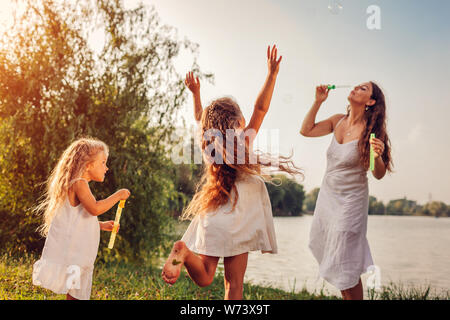 Madre aiuta le figlie a soffiare bolle nel parco d'estate. I bambini divertendosi e cattura bolle all'esterno. Famiglia di trascorrere del tempo insieme Foto Stock