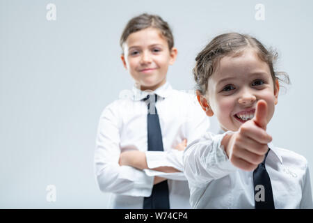 Felice motivati bambina dando un pollice in alto con la mano tesa per la telecamera e un raggiante sorriso amichevole come egli sorella guardato da dietro wi Foto Stock