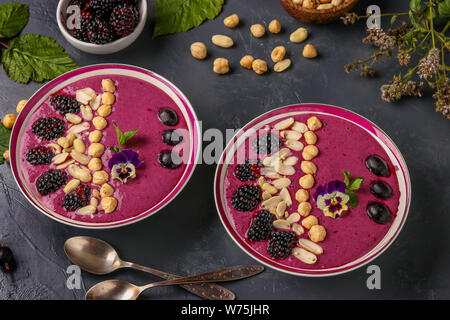 Sana colazione smoothie ciotola con more, ribes, arachidi, nocciole, decorato con fiori su sfondo scuro, la foto in orizzontale Foto Stock