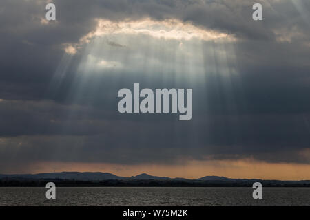 Sunray a vicino al tramonto con nuvole scure sullo sfondo di un cielo arancione e il lago Trasimeno (Umbria, Italia) al di sotto di Foto Stock