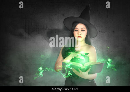 Asian witch woman in hat apprende l'incantesimo dal libro magico con un muro nero lo sfondo. Concetto di Halloween Foto Stock