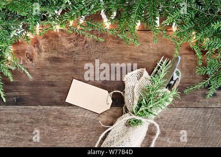 Albero di natale rami di pino, argenteria avvolta in una tela igienico con la scheda vuota e luci decorative su un sfondo rustico di legno del granaio. Immagine Foto Stock