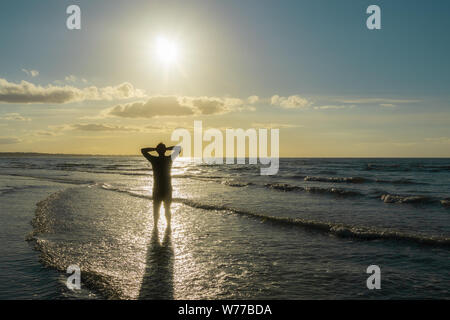 Silhouette di un giovane uomo in piedi sulla riva del mare di fronte al sole. All'aperto, estate, libertà concetto con copia spazio. Foto Stock