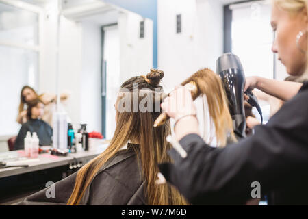 Ritratto di donna felice presso il salone di parrucchiere. Professional hair styling concept. Parrucchiere ragazza di asciugatura dei capelli lunghi con phon e spazzola. Asciugatura con Foto Stock