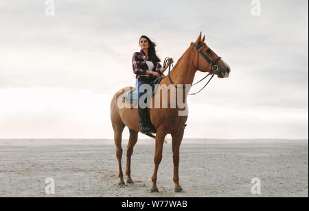 Per tutta la lunghezza della giovane donna seduta sul suo stallone presso la spiaggia. Equestre femmina sul suo cavallo in piedi presso la spiaggia. Foto Stock