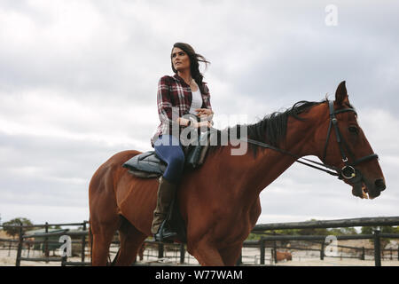 Giovane donna seduta sul suo cavallo e guardando lontano. Femmina di equitazione jockey il suo cavallo nel recinto. Foto Stock