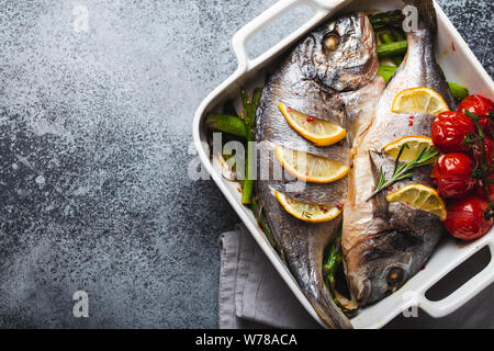 Pesce al forno dorado con asparagi verdi e pomodori in ceramica bianca teglia sul grigio cemento rustico sfondo, vista dall'alto. Una sana cena a base di pesce Foto Stock