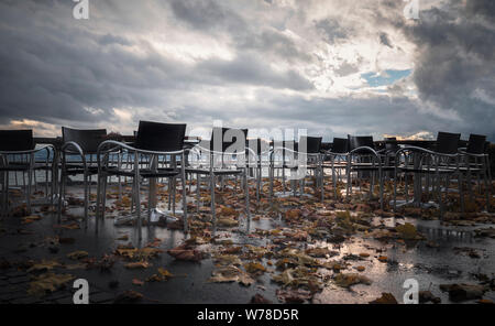 Cupo autunno con arancia caduta foglie tra tabelle vuote e sedie da bagnato su una veranda, in una piovosa giornata di novembre, sul lago di Costanza, in Germania. Foto Stock