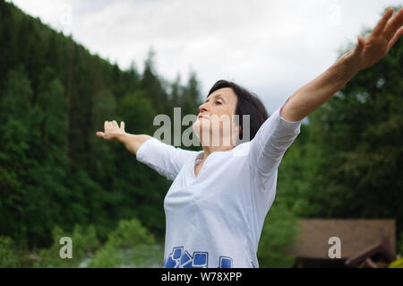 Senior donna facendo un esercizio di stretching per la parte superiore delle braccia al di fuori oltre il paesaggio di boschi e montagne Foto Stock