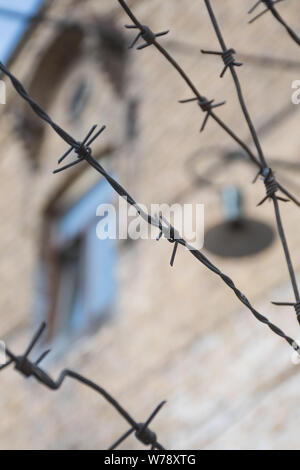 Close-up di un arrugginito del filo spinato che circonda una concentrazione e sterminio camp, vecchia baracca con finestra e lampada in soft focus Foto Stock