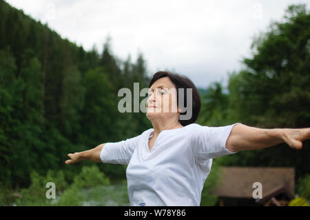 Senior donna facendo un esercizio di stretching per la parte superiore delle braccia al di fuori oltre il paesaggio di boschi e montagne Foto Stock