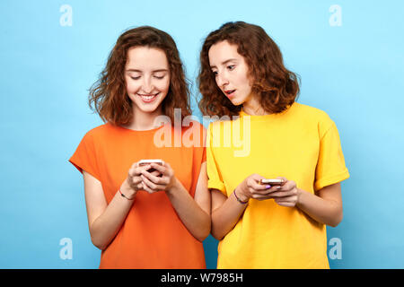 Due gemelli tenendo i telefoni cellulari, effettuando una chiamata telefonica, giocando i giochi online, la navigazione, la navigazione in rete . isolato sfondo blu, studio sho Foto Stock