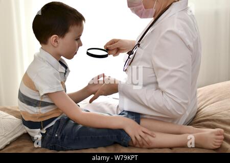 Un medico di famiglia in un accappatoio siede su un letto esamina i microbi sulle palme del bambino attraverso una lente di ingrandimento con una penna. Foto Stock