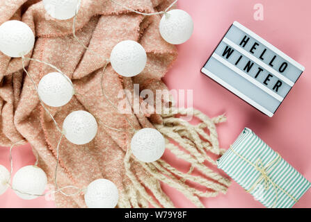 Concetto di Natale lay piatto. Caldo e accogliente bianco inverno maglione abbigliamento, Lightbox e decorazioni di Natale cornice rosa su sfondo pastello Foto Stock