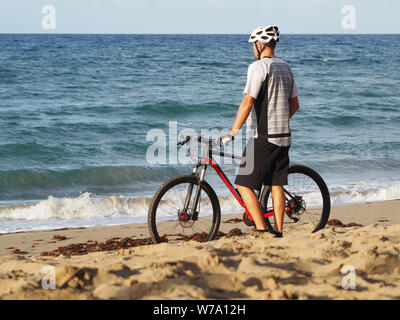 Ciclista in appoggio sulla spiaggia dell'Oceano Atlantico guarda verso l'oceano. Vista posteriore. Tramonto di sera. Foto Stock