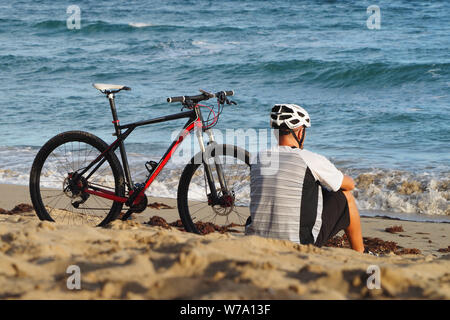 Un uomo con una bicicletta è in appoggio sulla spiaggia dell'Oceano Atlantico. Si siede nella sabbia. Vista posteriore. Foto Stock