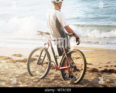 Ciclista in appoggio sulla spiaggia dell'Oceano Atlantico. Vista posteriore. Tramonto. sun flare. Foto Stock