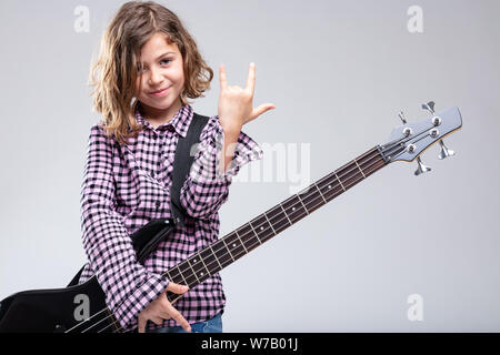 Sorridente ragazza giovane a suonare la chitarra dando un segno di corna gesto con la sua mano per il rock e musica heavy metal su uno sfondo grigio con spazio di copia Foto Stock