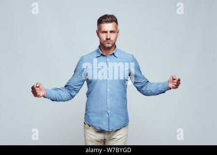 Sono forte. Uomo con bearded sicuro e bello in abiti casual che tengono le mani aggrappate in pugni mentre si levano in piedi contro sfondo grigio. Forza e motivazione. Scatto in studio Foto Stock