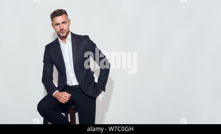Intelligente e sicuro. Ritratto di affascinante uomo d'affari con bearded in costume classico guardando la macchina fotografica mentre si siede sulla sedia su sfondo grigio. Concetto di successo. Uomini bellezza. Scatto in studio Foto Stock