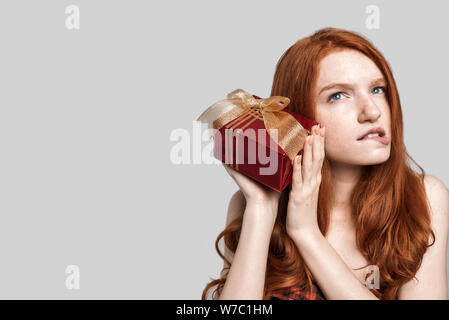 Studio shot di carino e incuriosito redhead donna tenendo un regalo vicino al suo orecchio e rendendo preoccupato faccia mentre in piedi contro lo sfondo grigio Foto Stock