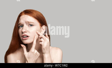 Studio shot di preoccupato giovane donna redhead esaminando il suo volto mentre in piedi contro uno sfondo grigio. L'Acne. Cura della Pelle Foto Stock