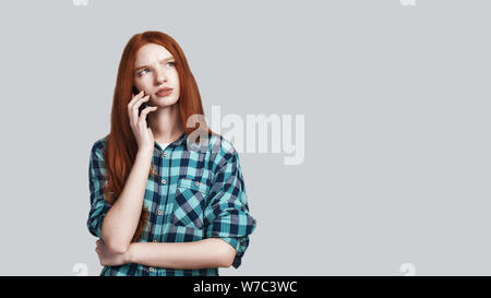 Studio shot di giovani redhead donna in abbigliamento casual sta parlando al telefono e rendendo il volto triste e stando in piedi contro uno sfondo grigio. Le emozioni negative. Digital Foto Stock