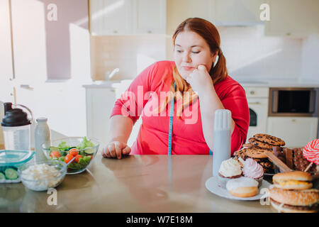 Fat giovane donna in cucina e salotto a mangiare cibo. Lei guarda pasto sano sul lato sinistro. Soft misurare il nastro attorno al collo. Dolce il cibo spazzatura di destra sul sid Foto Stock