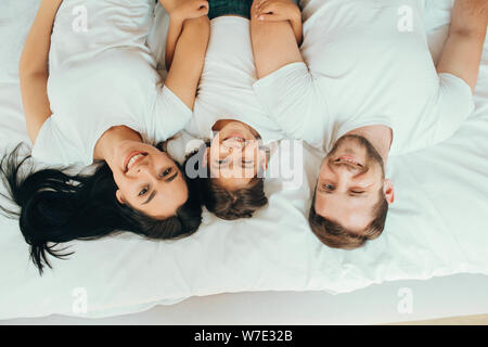 La famiglia felice giacente su un nuovo letto bianco abbracciando e guardando la telecamera, vista dall'alto. Madre, padre e figlia insieme giacente sul letto di casa Foto Stock
