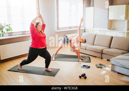 Giovani slim e modelli in sovrappeso exersicing insieme in salotto. In piedi di asana o yoga pone. Allenamento per due. Manubri sul pavimento Foto Stock