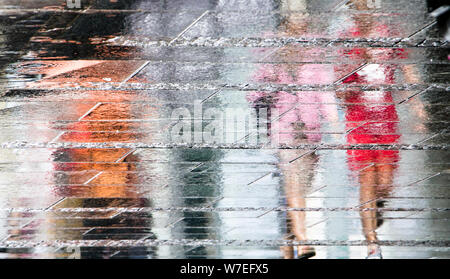 Sfocata riflessione ombra sagome delle persone che camminano in una piovosa città pedonale strada bagnata in un giorno di estate in una pozza Foto Stock