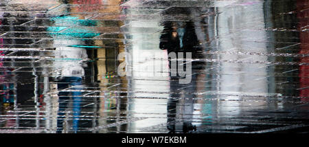 Sfocata riflessione ombra sagome delle persone che camminano sotto ombrellone in una piovosa città pedonale strada bagnata in una pozza Foto Stock