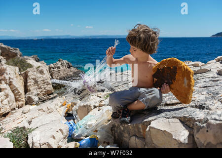 Little Boy pulizia spiaggia piena di bottiglie in plastica rigida e altra immondizia lavato fuori sulla costa. Foto Stock