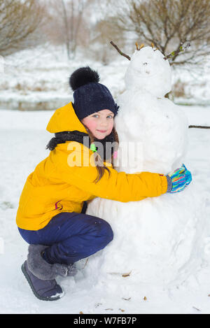 Ritratto di bella bambina in inverno in giacca gialla e blu hat. bambino felice fa un pupazzo di neve. Foto Stock