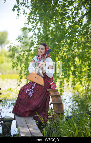 Giovane donna sorridente in tradizionali abiti russo si siede sul piccolo ponte vicino al lago e tenendo la balalaika Foto Stock