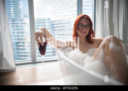 Un ginger donna sorridente giacente nel bagno schiuma e tenere un bicchiere di vino rosso - una vista su un vetro di moderni edifici dalle finestre panoramiche Foto Stock