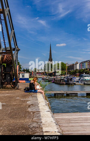 Due persone sedute al sole con i libri sulla banchina del porto di galleggiante con St Mary Redcliffe in background, Bristol, Regno Unito. Luglio 2019. Foto Stock