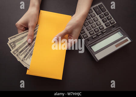 Close-up. La ragazza è in possesso di una busta gialla con il denaro su un cuoio scuro sfondo, sul tavolo è una calcolatrice. Stipendio o corrompere concept Foto Stock