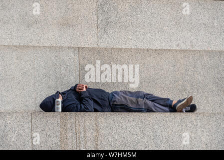 Bianco maschio caucasico in felpa con cappuccio con cappuccio sulla faccia e la birra lager può dormire sul plinto passi della Colonna di Nelson, in Trafalgar Square, Londra, Regno Unito. Foto Stock