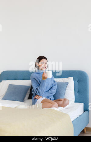 Ragazza in un maglione blu in interni in stile Hygge con un bicchiere in mano si siede sul letto Foto Stock