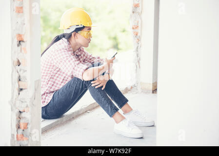 Giovane lavoratore edile azienda smart phone e sittin al sito in costruzione.giovane bella ingegnere di lavoro tenendo i progetti di un edificio giallo usura timone Foto Stock