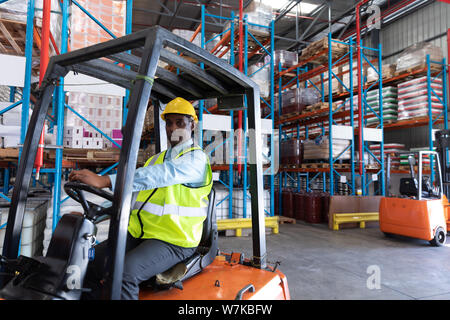 Lavoratore di sesso maschile guardando la fotocamera mentre si guida il carrello elevatore in magazzino Foto Stock