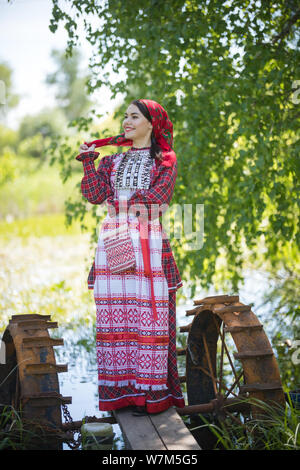 Donna attraente in sciarpa e tradizionale russo vestiti sorge su un piccolo molo vicino al lago, guarda a destra, che posano per una foto Foto Stock