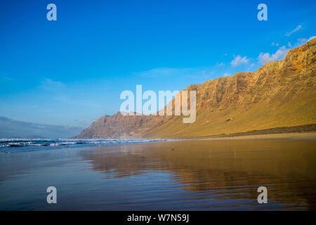 Spagna, Lanzarote, popolare spiaggia di surf Famara riflettendo impressionanti montagne di famara Foto Stock