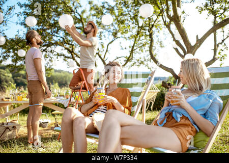 Giovani fidanzate rilassarsi sui lettini con ragazzi decorare giardino no lo sfondo, la preparazione per la festa durante la giornata di sole Foto Stock