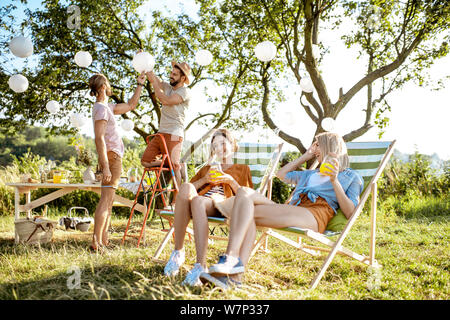 Giovani fidanzate rilassarsi sui lettini con ragazzi decorare giardino no lo sfondo, la preparazione per la festa durante la giornata di sole Foto Stock