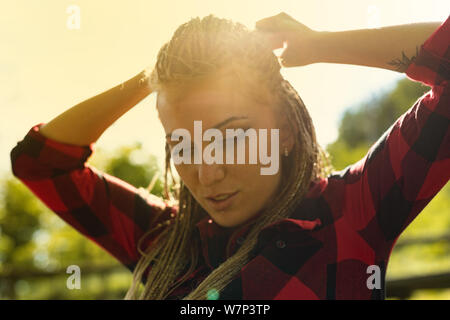 Donna adulta in maglietta rossa legatura di capelli dietro la sua testa con il verde degli alberi e il campo in background sotto il sole luminoso Foto Stock