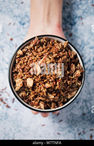 Elevato angolo di visione della mano di un uomo caucasico tenendo un bianco di ceramica ciotola con fritti riso rosso con dadolata di carne di pollo, su un tavolo di marmo o un contatore Foto Stock