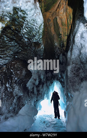 Uomo in piedi alla bocca della caverna riempita di ghiaccioli / stalattiti di ghiaccio, il lago Baikal, Siberia, Russia, Marzo. Foto Stock