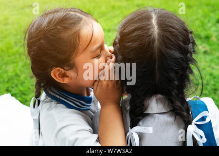 Due giovani ragazze whispering e condividere un segreto durante la sessione di giochi sul verde sullo sfondo di vetro Foto Stock